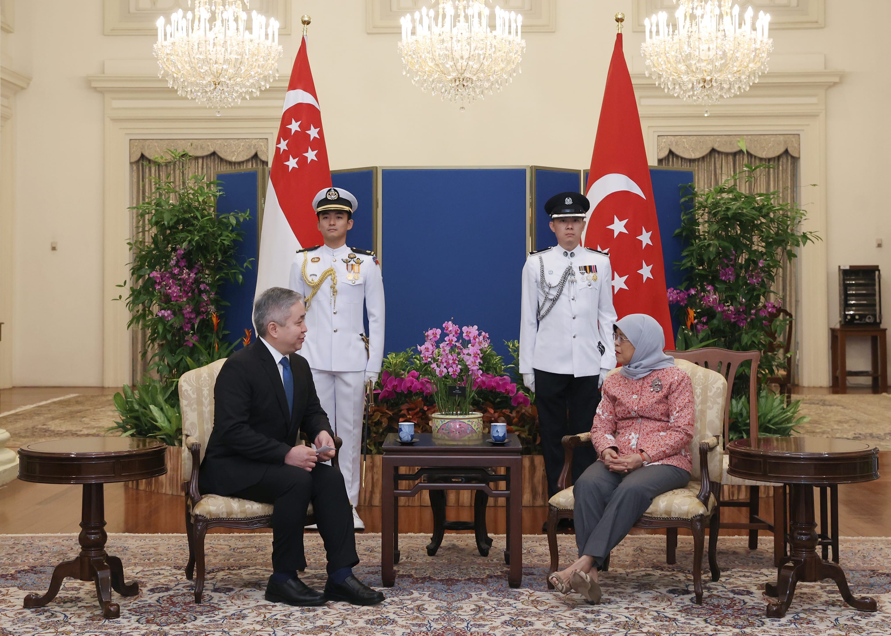 Two people sit facing each other, guarded by two standing figures by Singapore flags.
